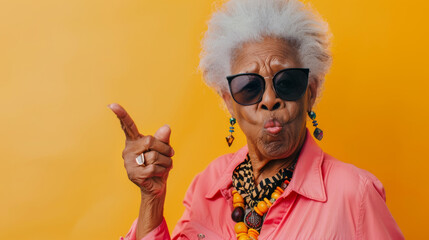 A spirited elderly woman with sunglasses pointing with panache against an orange backdrop. - obrazy, fototapety, plakaty