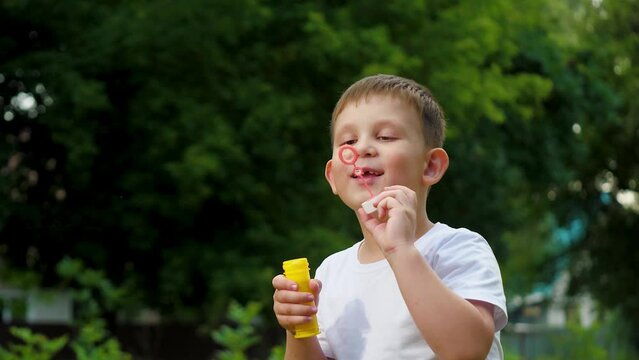 Little joyful boy in t-shirt blows soap bubbles playing in summer city park. Cheerful boy blows bubbles in park among trees on vacation. Schoolboy spends time blowing soap bubbles slow motion