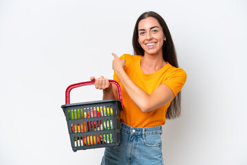 Young caucasian woman holding a shopping basket full of food isolated on white background pointing back