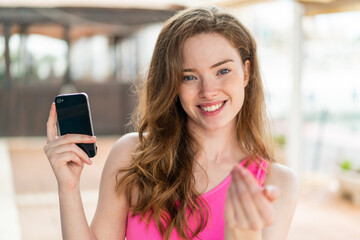 Fototapeta na wymiar Young redhead woman using mobile phone at outdoors inviting to come with hand. Happy that you came
