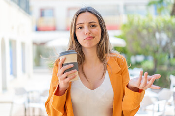 Young pretty woman holding a take away coffee at outdoors making doubts gesture while lifting the...