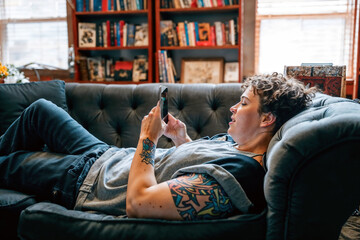 LGBTQ+ person lounging on a sofa, comfortably using a smartphone, depicting everyday life and modern connectivity.

 - obrazy, fototapety, plakaty
