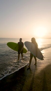 Vertical video of two dark-skinned young men walking along the seashore dressed in wetsuits and carrying surfboards. African surfing couples. Water sports. Young people look at each other at sunset.