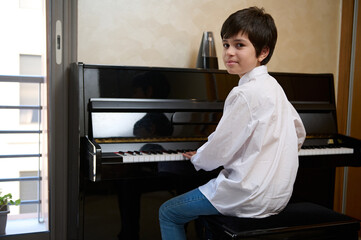 Happy teen boy in white casual shirt, sitting by piano and smiling at camera while performing...