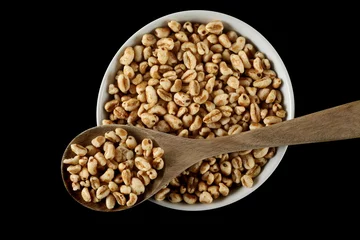 Poster Puffed wheat cereal flakes in wooden spoon and bowl isolated on black, top view © dule964