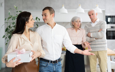 Positive interested young adult man gesturing welcomingly towards female guest holding gift box, against backdrop of disapproving elderly parents in domestic setting.... - Powered by Adobe