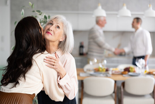 Hospitable friendly elderly woman warmly greeting and kissing daughter-in-law coming for cozy family get-together with set table in background