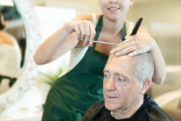 Senior gray-haired male client getting stylish haircut in hairdressing salon from skillful female...