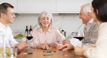 Mature friends is playing card game poker with middle-aged spouses. Married couple pensioners have fun during meeting and gatherings, enthusiastically excitedly fulfill conditions of game with friends