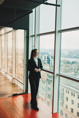 A modern smiling business woman in a black suit and glasses is standing in the office near the window. Woman manager in glasses and white shirt.