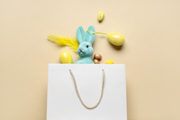 Shopping bag with toy bunny, Easter eggs and feather on beige background