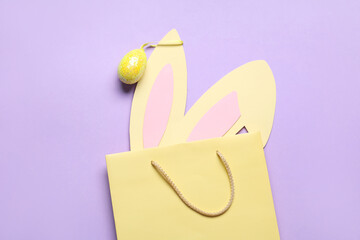 Shopping bag with paper Easter bunny ears and egg on lilac background