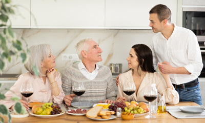 Senior couple gathered at kitchen table with grown son and daughter-in-law, enjoying conversation with wine and light snacks..
