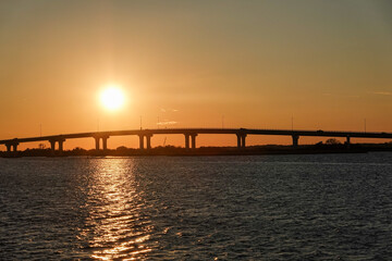 Fototapeta na wymiar Sunset over a long bridge with an orange sky and reflection on the bay water