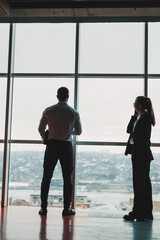 A man and a woman are standing in the office against the background of a large panoramic window. Modern spacious workspace with employees. Business partners