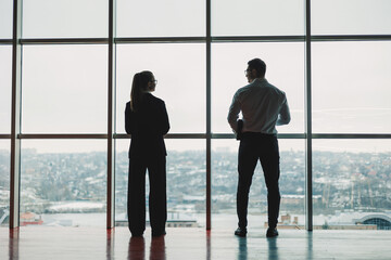 Fototapeta na wymiar A man and a woman are standing in the office against the background of a large panoramic window. Modern spacious workspace with employees. Business partners