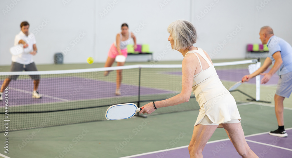 Wall mural Rear view of fit elderly woman playing pickleball on indoor court, swinging paddle to return ball over net ..