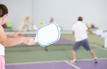 White paddle-shaped racquet for pickleball in hand of young female player swinging to hit ball during friendly match in close court, selective focus