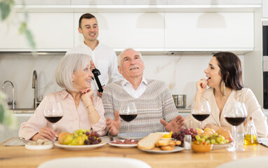 Mature couple are sitting at table, chatting sweetly, discussing and telling news, sharing plans with adult children. Senior woman and man celebrate housewarming party with middle-aged guests spouses