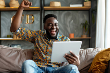 Man with tablet celebrating success. A radiant young black man exudes sheer happiness as he raises his fist in a triumphant gesture, holding a tablet in his other hand. - obrazy, fototapety, plakaty