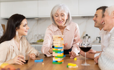 Obraz na płótnie Canvas Cheerful senior woman spending free time with husband and adult children at laid-back family home get-together with drinks, concentrating on removing block from tower while playing jenga
