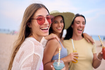 Portrait of cheerful three young Caucasian women posing smiling looking at camera on beach. Happy friends of generation z holding fruit smoothies enjoying their summer holidays in Mediterranean sea