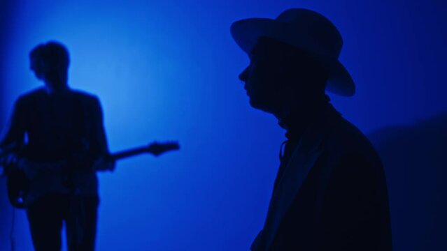 Side tilt footage of silhouettes of keyboardist singing and playing synthesizer and guitarist accompanying in studio with neon blue color filter