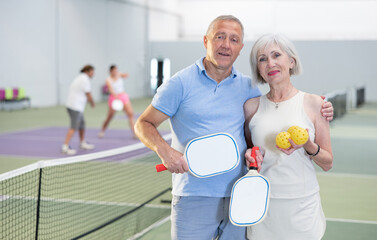 Happy smiling elderly couple, man and woman in sportswear with rackets and balls in hands posing near net on indoor pickleball court after friendly match