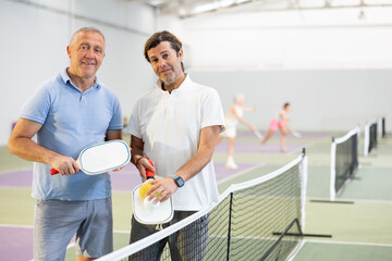 Two positive smiling men in sportswear with rackets and balls in hands posing near net on indoor pickleball court after friendly match..