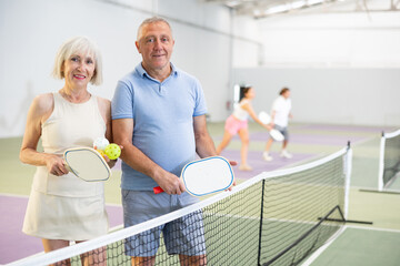 Happy smiling elderly couple, man and woman in sportswear with rackets and balls in hands posing near net on indoor pickleball court after friendly match - Powered by Adobe