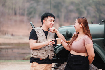 Cheerful couple enjoys a lighthearted conversation with warm drinks in hand, standing by an off-road vehicle in a natural setting.