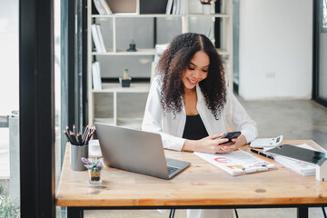 Smiling businesswoman multitasking, using a smartphone and laptop at her organized work desk in a contemporary office.