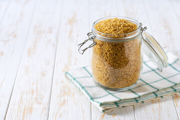Raw bulgur in glass storage jar on a white wooden table.