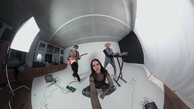 Caucasian female vocalist of glam rock band filming process of recording music video with musicians in professional studio with white background using action camera 360