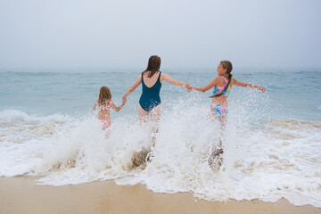 Mother and children playing on the ocean beach. Family enjoying the ocean. Mother holds girls's hands and they all look at the ocean together.