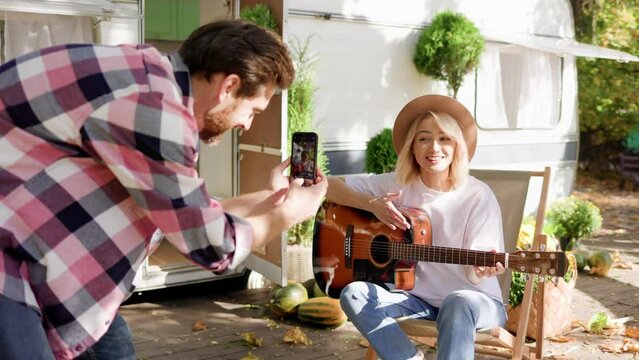 Funny and cheerful young couple. The girl plays the guitar, the guy photographs her for social networks against the backdrop of a travel trailer. Weekends in the summer time. Romance in nature.
