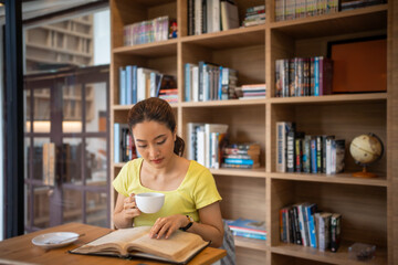 Young woman reading book and drink hot coffee in library at home, smiling, laughing, enjoying...