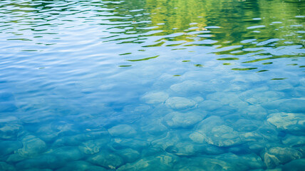 The shallow water of a lake with faint ripples on the surface and smooth rocks; a reflection of trees is shown.
