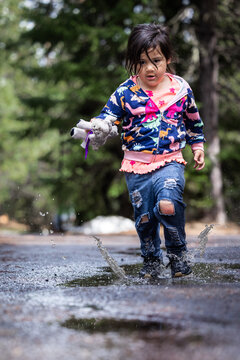 A little girl is running through a puddle of water, holding a stuffed animal