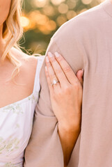 An engaged woman has her hand with engagement ring looped around her fiance's arm at sunset. 