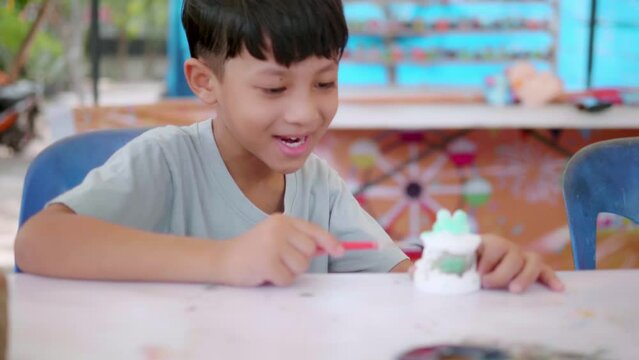 Cute Asian boy holding a paintbrush is painting watercolors on various shapes with preschool children, art class participants. Developing children's abilities, creative hobbies, day care activities