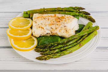 Cod fillet baked with garlic butter sauce served with fresh spinach and roasted asparagus on a white plate close-up