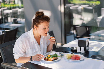 A woman in a white shirt eats lunch or breakfast outdoors in a cafe.