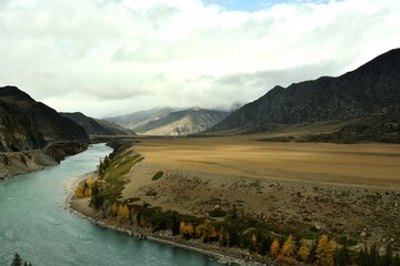 A turn in the course of a wide turquoise river flowing through a huge valley in the mountains on a cloudy autumn day.