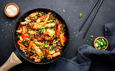 Hot stir fry chicken  slices with red paprika, mushrooms, chives and sesame seeds in frying pan. Black table background, top view