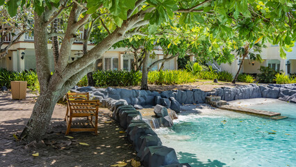 A relaxation area by the swimming pool with turquoise water. A wooden bench stands in the shade...