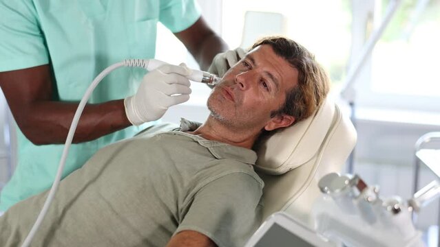 European man lying on couch during skin purificating procedure with air hydrogen-oxygen cleaning mixture using multifunctional beauty machine