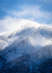 A closeup view of a mountain peak covered in snow as the wind swirls clouds around it. Aspen Springs, California - Highway 395