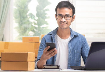 Asia young man smiling and happy holding smartphone looking at camera with parcel box on table and...