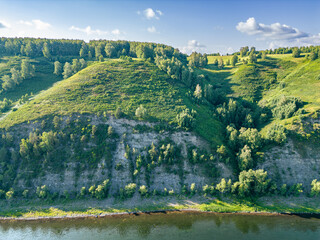 The steep rocky bank of the river is overgrown with forest, the roots of trees and bushes are...